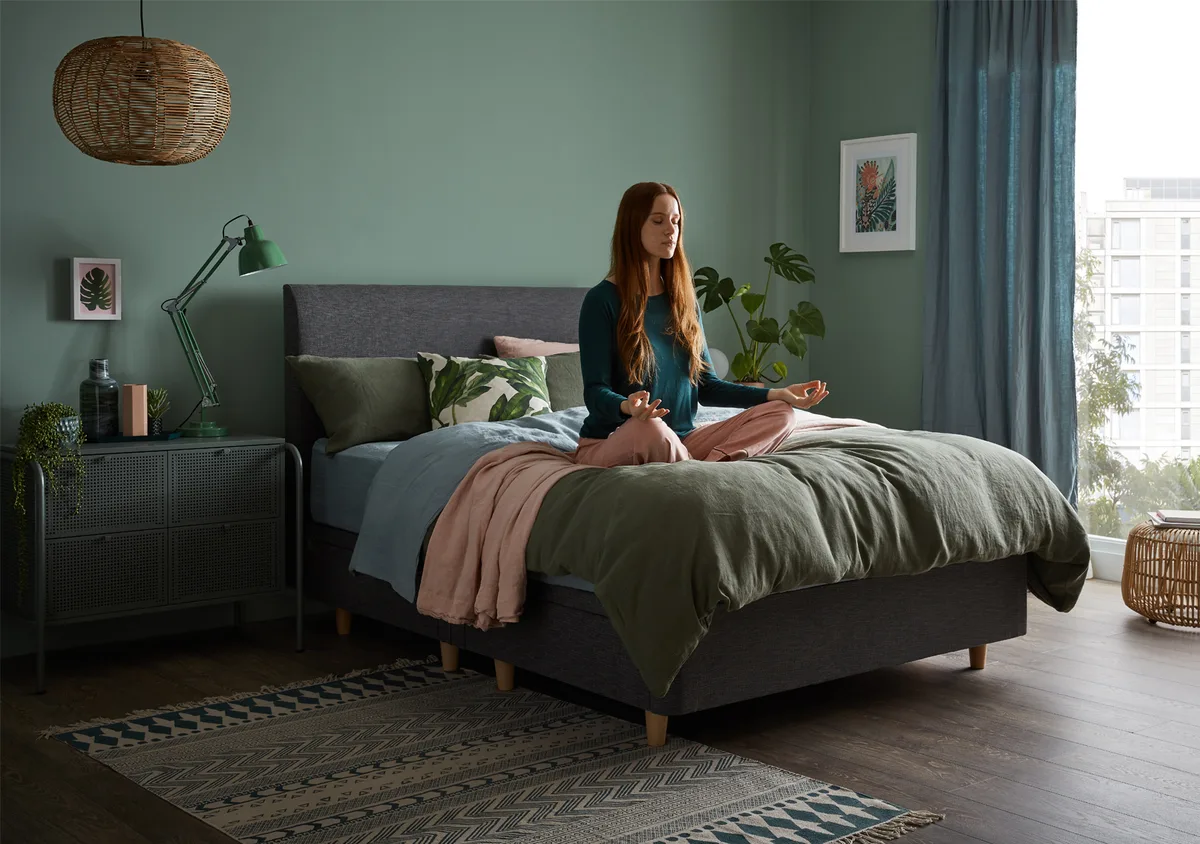 Young woman sitting cross-legged with her eyes closed on a double bed in a meditation pose