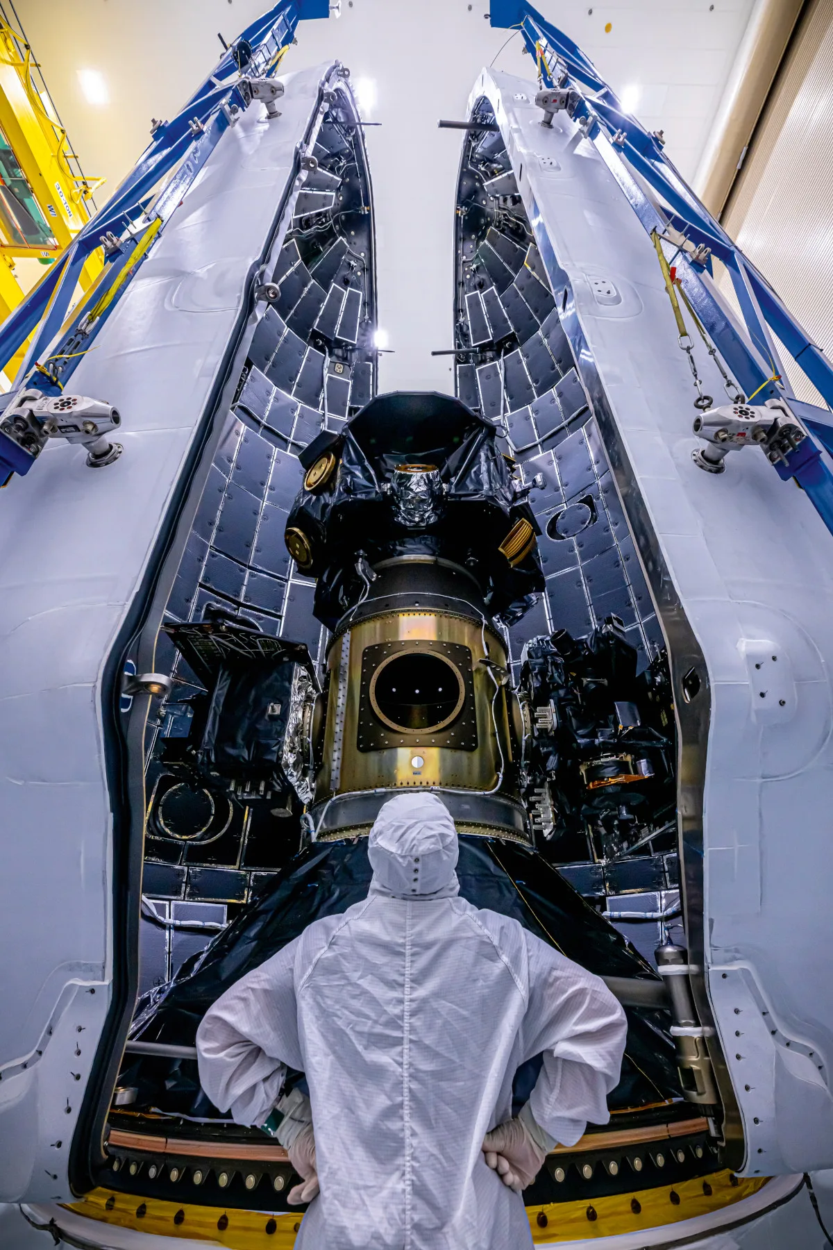 A scientist stands looking at the IMAP being loaded into the SpaceX Falcon 9 payload container. They have their hands on their hips and are wearing protective gear
