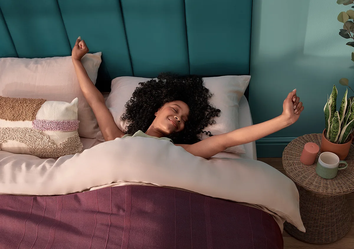 Smiling young woman in bed with dark curly hair spread across the pillow, eyes closed, arms stretching like she just woke up